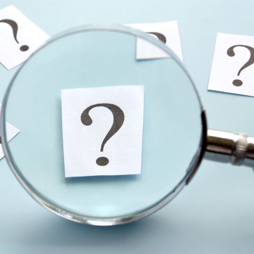 Magnifying glass with question mark in view on light blue background with other question marks on table-like background.