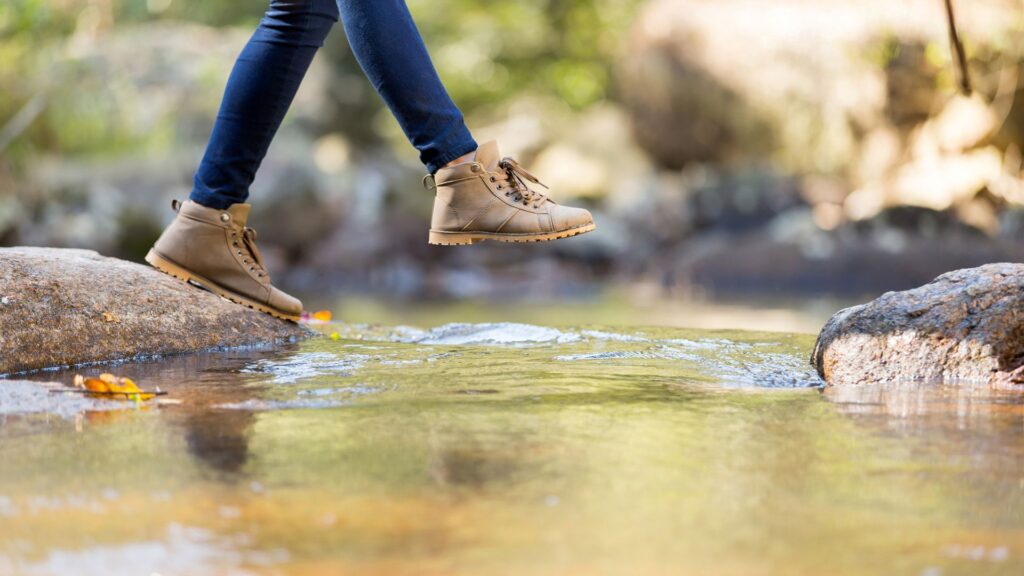 Woman in jeans and hiking boots stepping from one rock to the next. The rock she's stepping towards makes it look like she might splash in the water before reaching it.