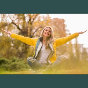 Woman sitting on top of mountain in grass, wearing a yellow sweater with a smile on her face and her arms outstreatched representing joy, accomplishment and success.