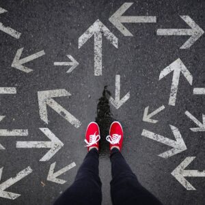 Woman wearing red sneakers and jeans stangin on asphalt road with arrows pointing in different directions all around her representing she doesn't know which way to go