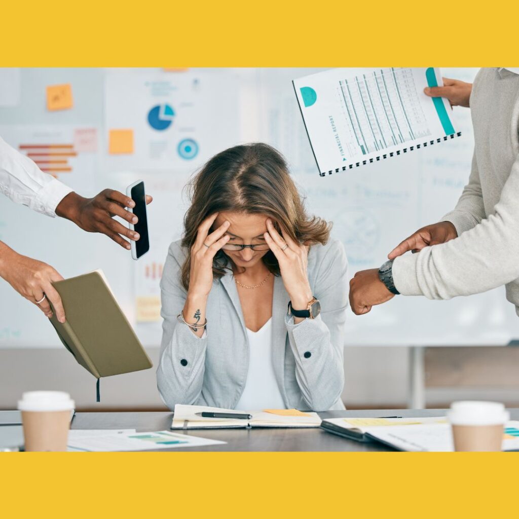 Woman sitting at desk holding her head in her hands bombarded by people attempting to get her attention